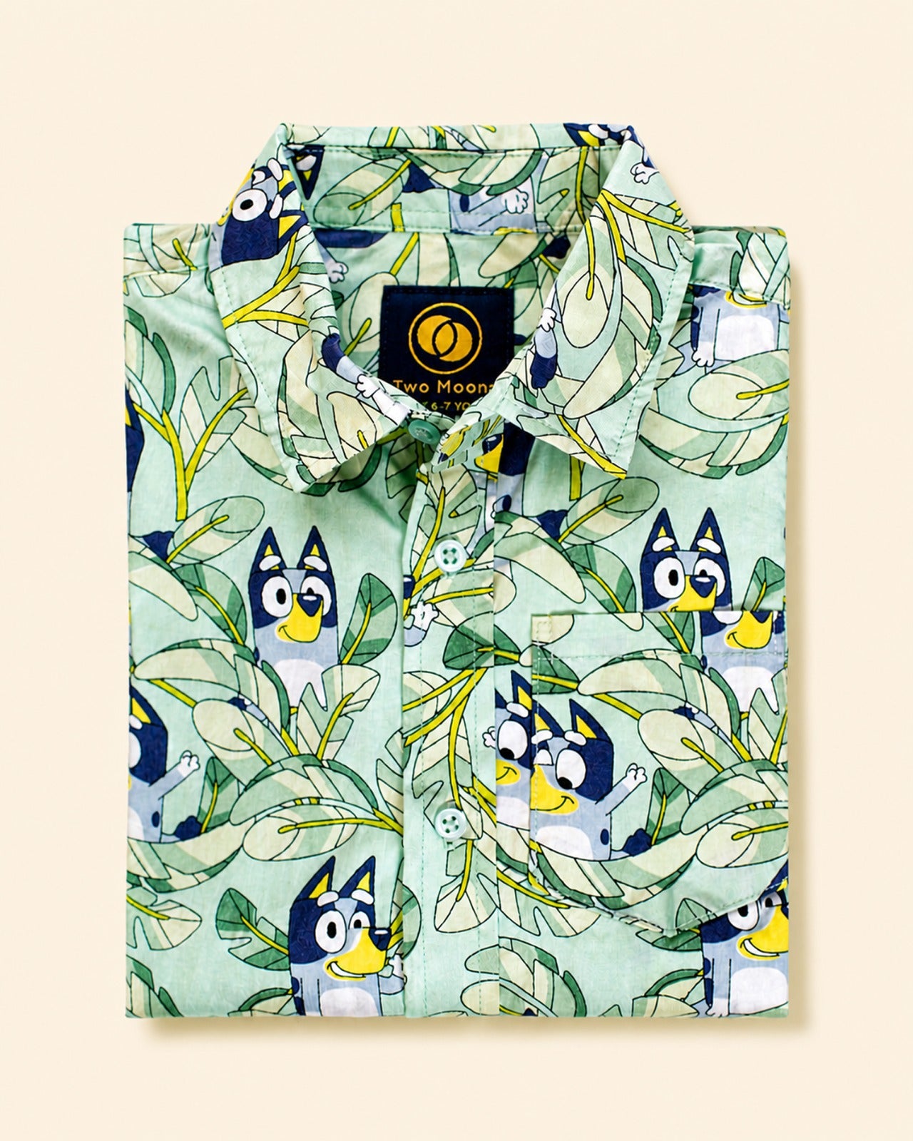 Folded Two Moons Canada kids short-sleeve button-down shirt featuring a cartoon blue dog and tropical green leaves on a mint background.